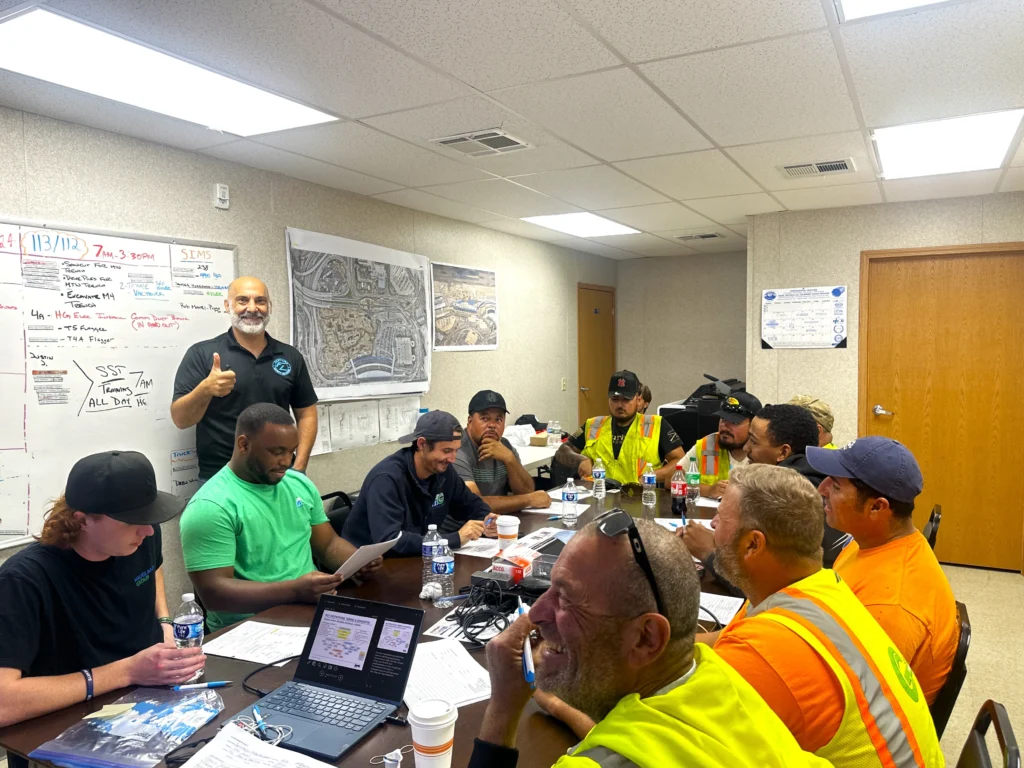 In a construction safety training class, a group of workers gather in a meeting room, surrounded by laptops, papers, and drinks. One person stands at the back, giving a thumbs-up as maps and notes on the walls highlight crucial safety protocols.