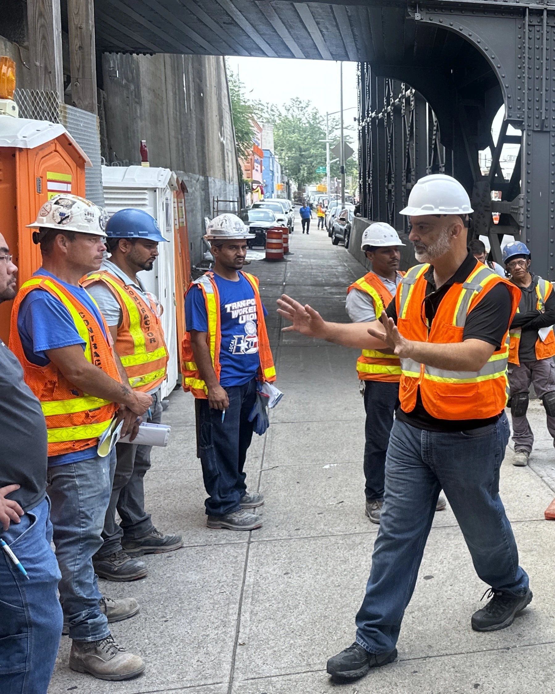 industrial workers attending a m.e.w.p. course by fast line safety instructor by concrete street in nyc. All students and instructors are wearing hard hat and high visibility orange vests.